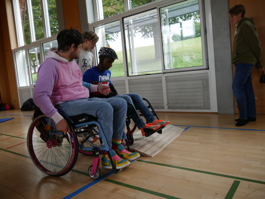 Hitzi, welcher im Rollstuhl ist, hilft den Kindern, die den Rollstuhl ausprobieren, wie man richtig über eine Rampe fährt.